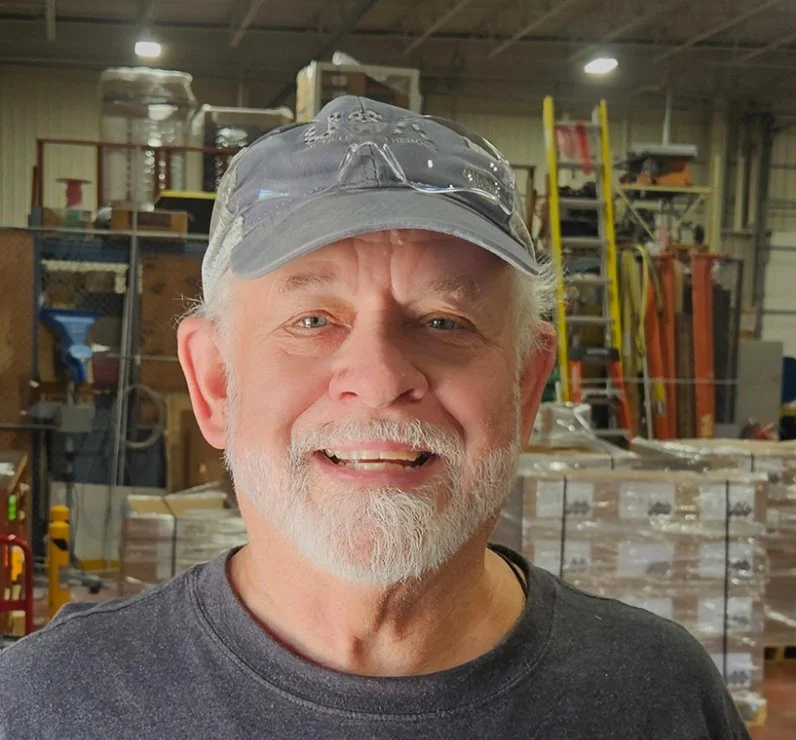 A smiling older man with a gray beard and mustache, wearing a baseball cap and safety glasses on top, standing inside a warehouse or workshop with shelves, ladders, and equipment in the background.