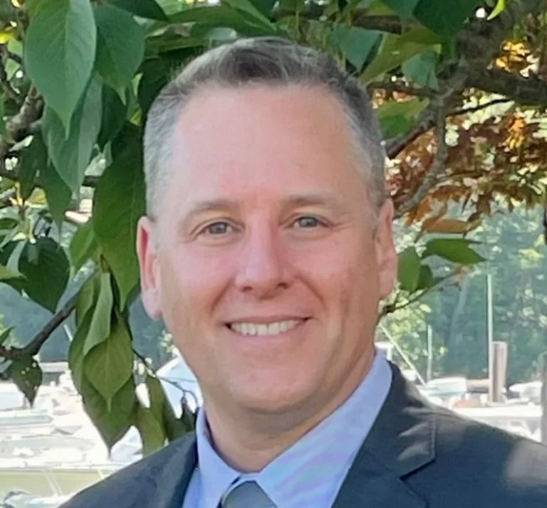 A man smiling outdoors with green leaves and trees in the background, dressed in a suit and tie.