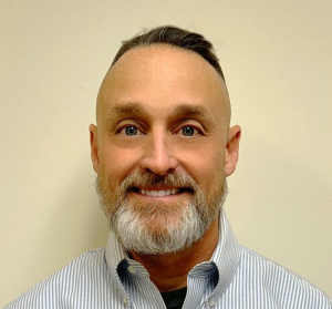Portrait of a man with a beard and short hair, wearing a collared shirt, smiling against a plain background.