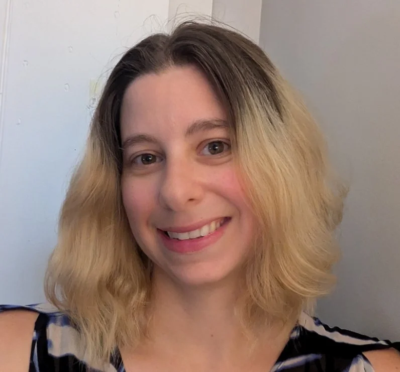 A woman with shoulder-length blonde hair smiling at the camera, wearing a sleeveless top with black and white stripes, against a plain light-colored background.