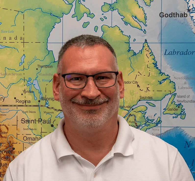 A smiling man with glasses and a white polo shirt stands in front of a map of North America, specifically Canada and the northern United States.