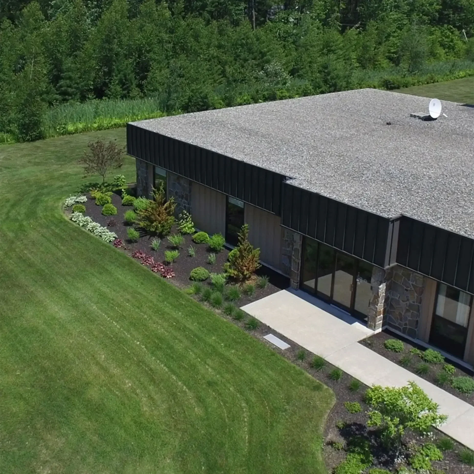 A black house with a flat, gravel roof, large window, and stone pillar exterior, surrounded by a well-manicured lawn with a flower bed, shrubs, and a walkway, with green trees and foliage in the background.