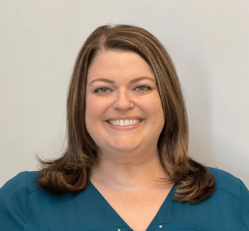 A woman with shoulder-length brown hair, blue eyes, and fair skin, smiling, wearing a teal top, standing against a plain light gray wall.