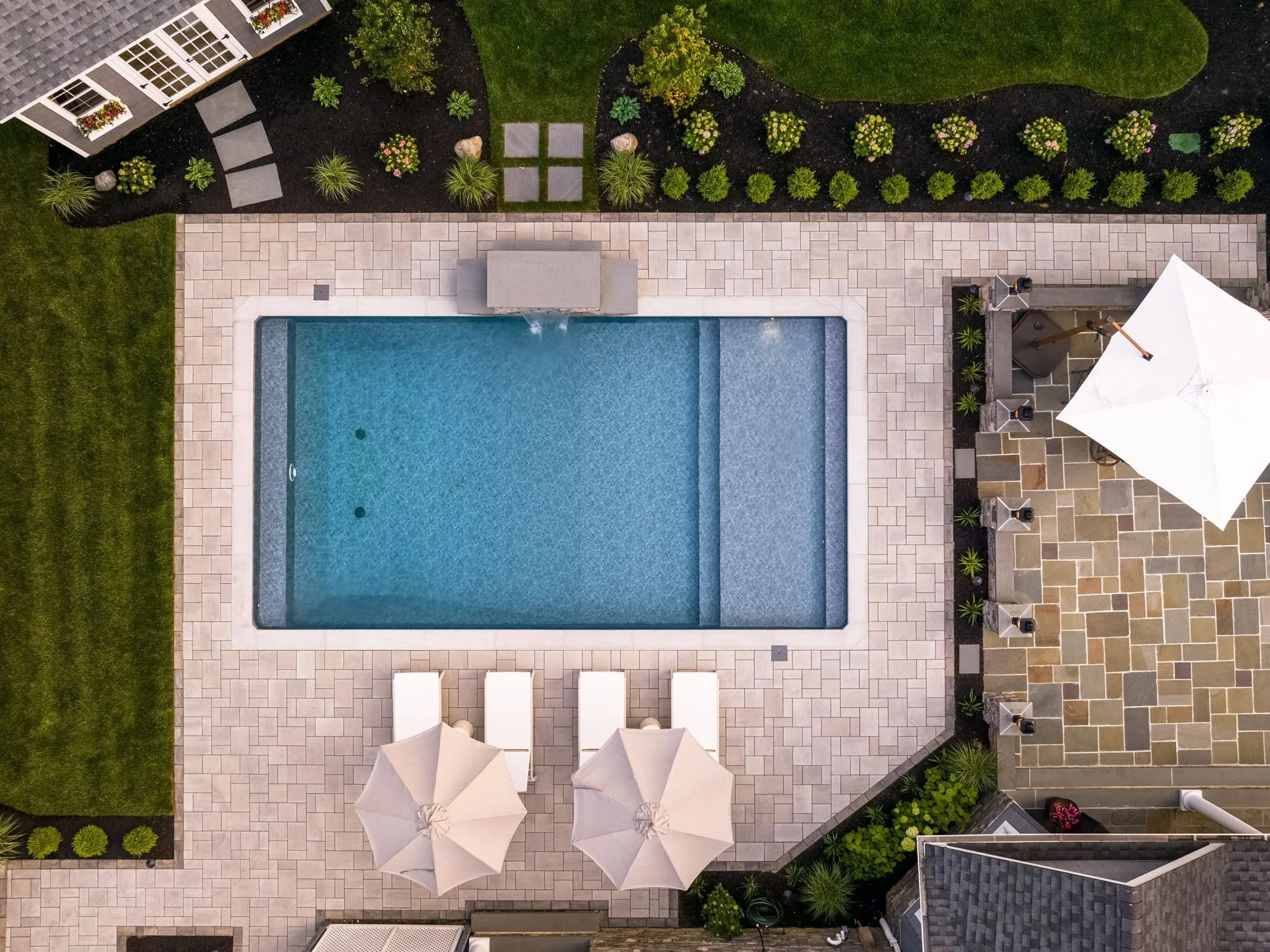 Aerial view of a backyard with a swimming pool, patio area with umbrellas and lounge chairs, a landscaped garden with plants and flowers, and part of a house with a covered porch.
