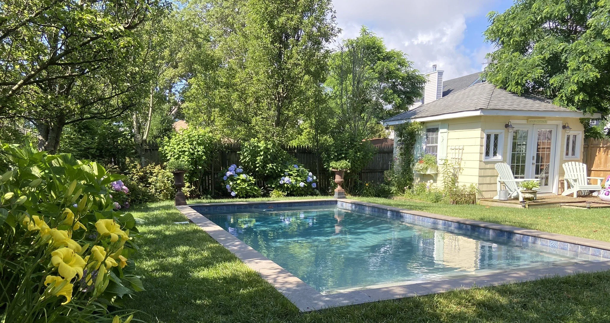 A backyard with a swimming pool, surrounded by green grass and lush trees. There are flowers, garden ornaments, and a small yellow house with white chairs outside.