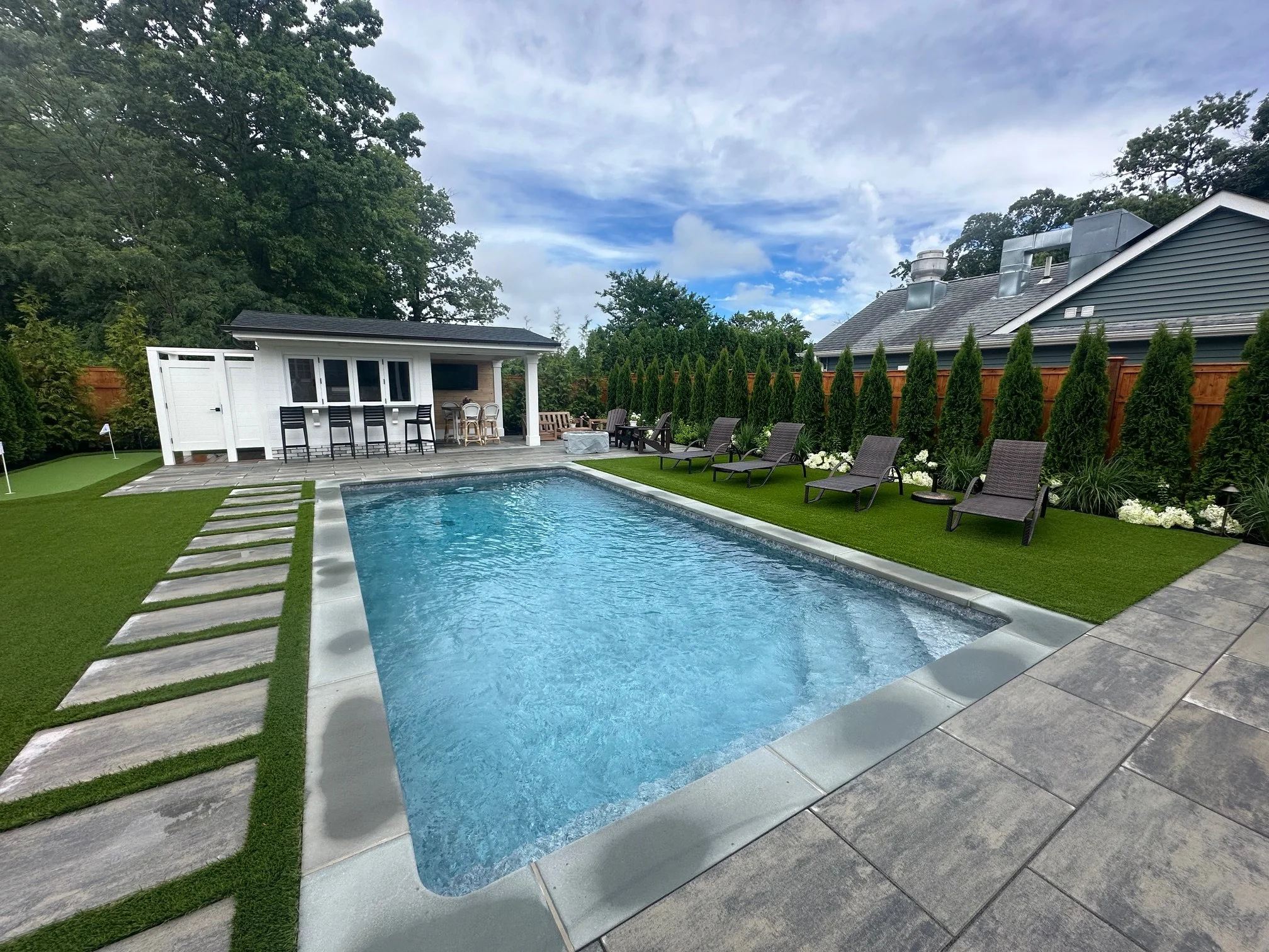 Backyard with swimming pool, lounge chairs, a pool house with barstools, surrounded by a wooden privacy fence and tall evergreen trees, with a cloudy sky overhead.