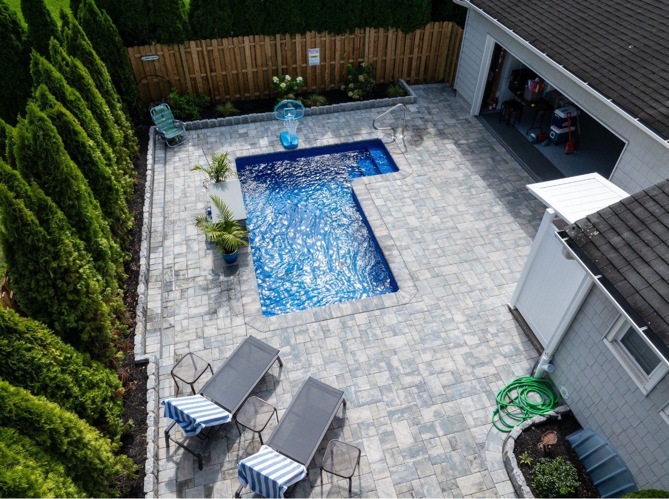 A backyard with a small swimming pool, surrounded by gray paver stones. There are two lounge chairs with striped towels, a small round table, a basketball hoop, and a few potted plants. The yard is enclosed by a wooden fence and tall hedge trees.