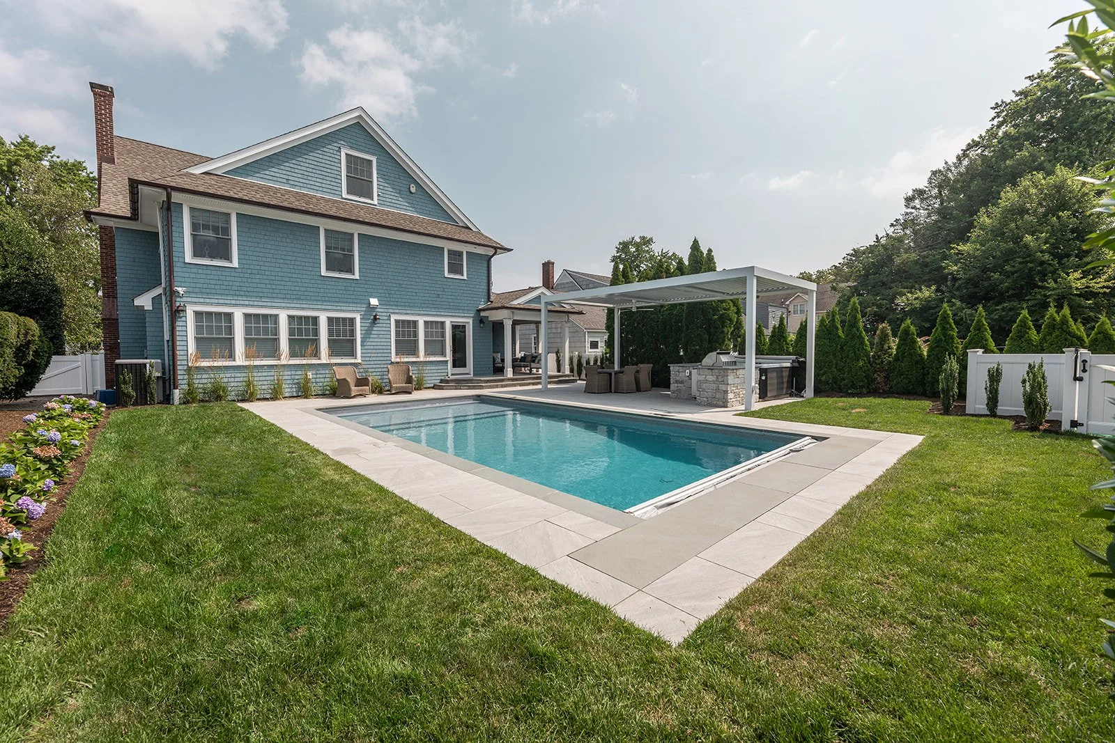 Backyard with a rectangular swimming pool, green lawn, blue house with multiple windows, outdoor seating area, and a shaded barbecue area.