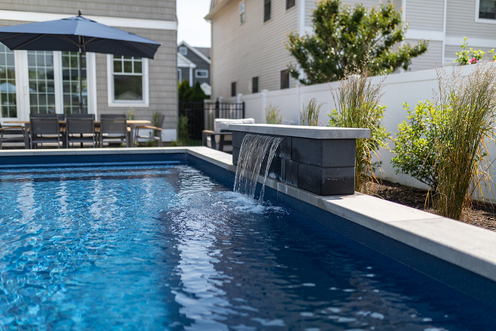  Backyard featuring a swimming pool with water feature, outdoor dining table with chairs, patio umbrella, and landscaped plants surrounded by white fencing and residential houses.