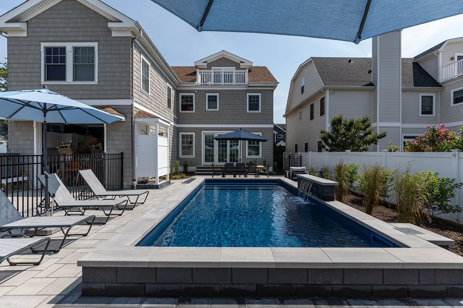 Backyard with swimming pool, lounge chairs, patio furniture, umbrellas, surrounded by white fence, with a gray house and neighboring house in the background.