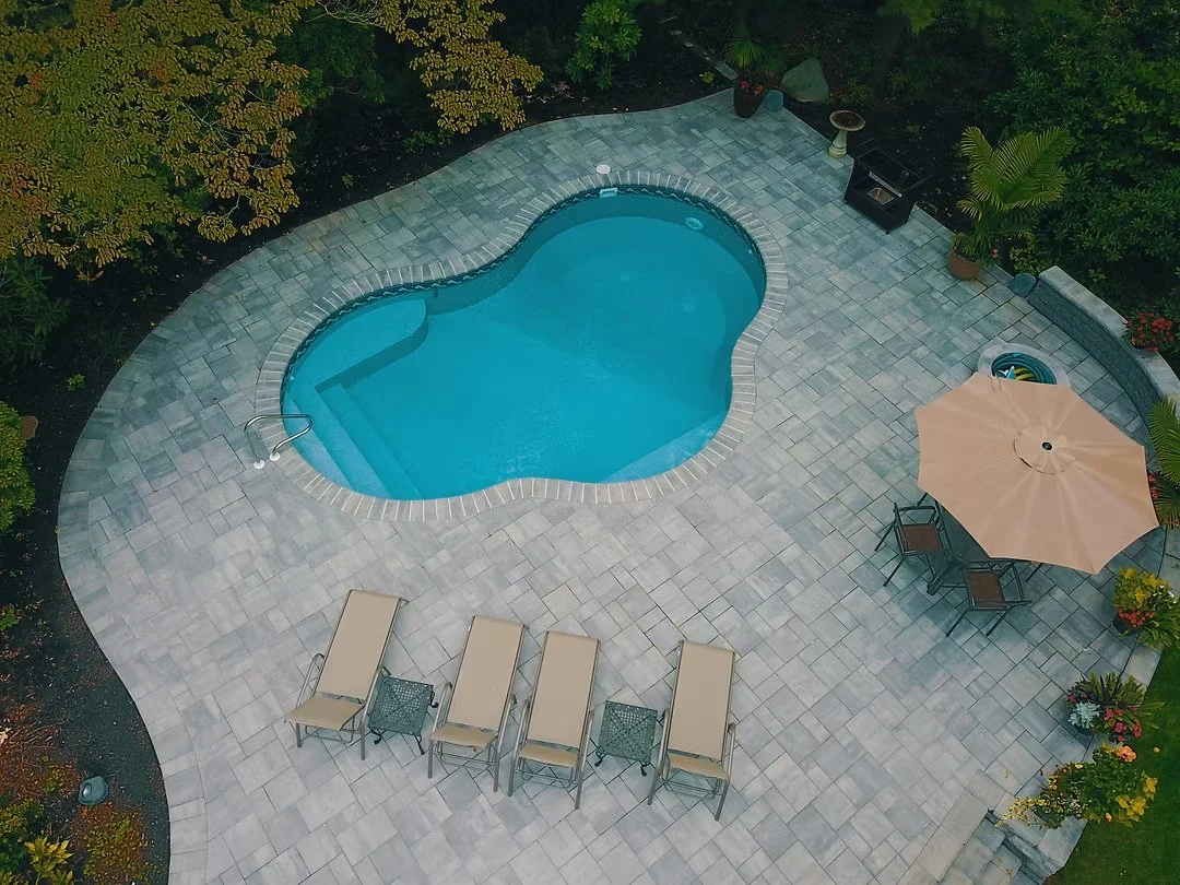 An aerial view of a backyard with a curved oval-shaped swimming pool surrounded by beige tiles. There are four beige lounge chairs, a side table, and a large beige umbrella with a set of chairs underneath. The yard is bordered by lush green trees and