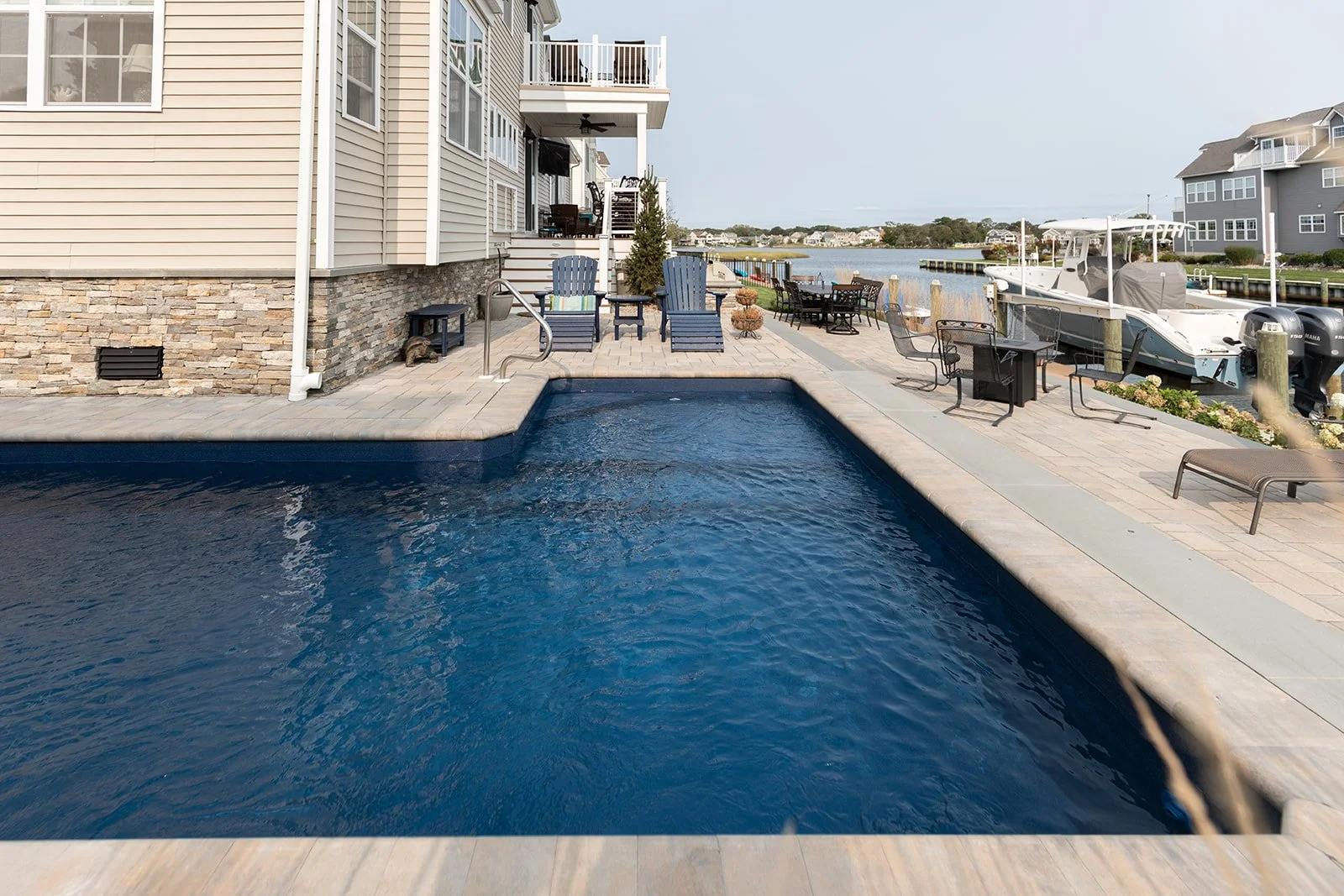 A backyard with a swimming pool, outdoor furniture, and a dock with boats on a waterway, with residential buildings in the background.