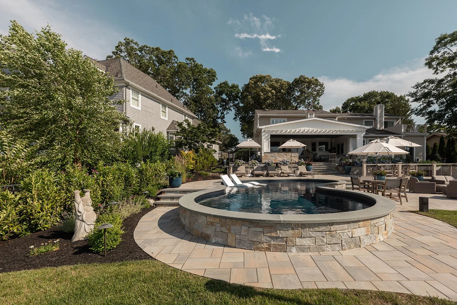 Backyard with a pool, surrounded by a patio with outdoor furniture, umbrellas, and lush greenery, with a large house in the background under a blue sky.