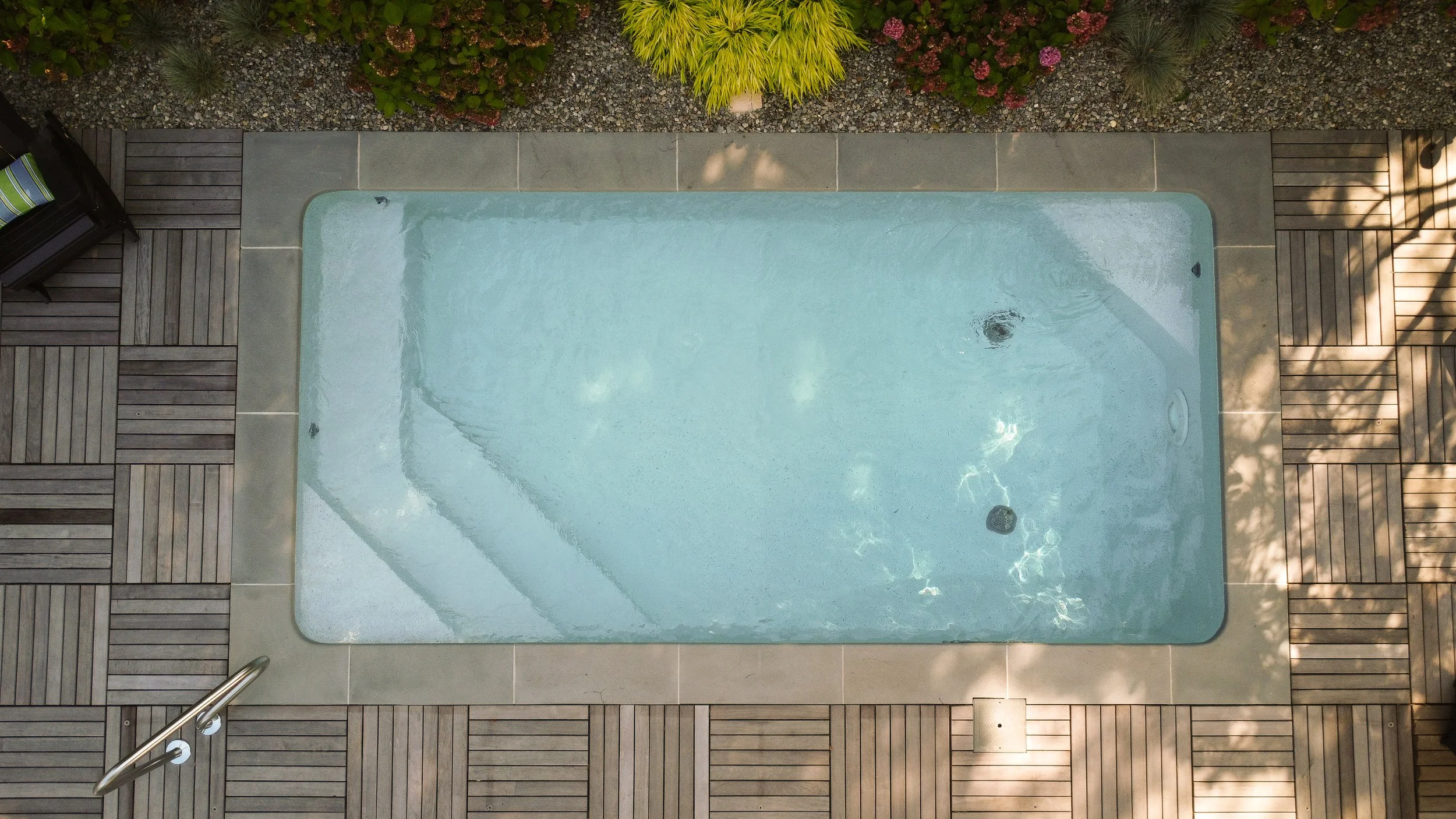Top-down view of a small rectangular swimming pool with clear water, surrounded by wooden decking and a gravel garden with bushes and plants.