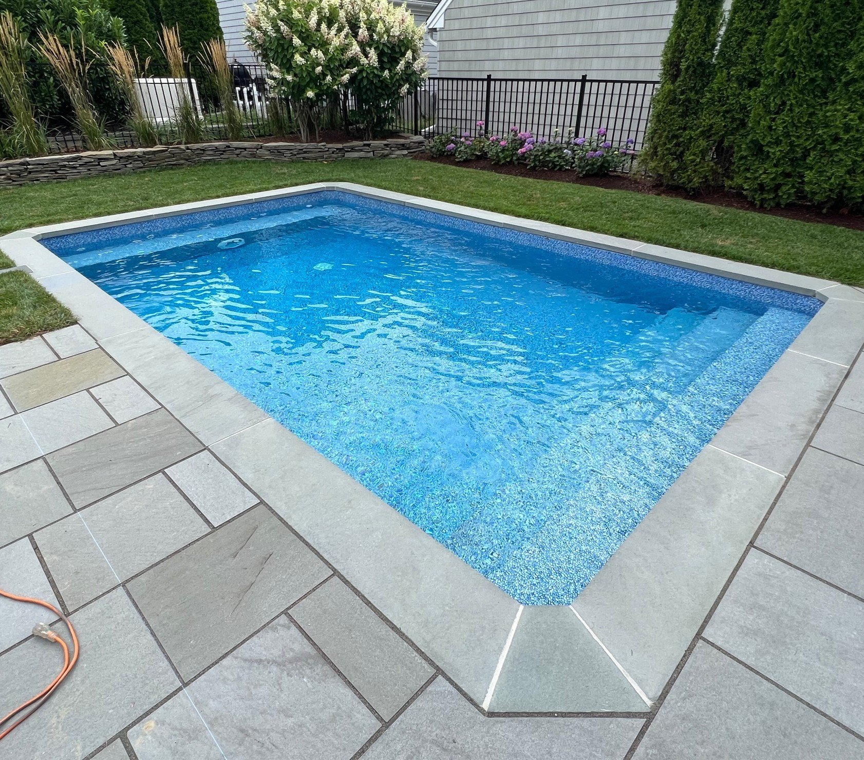 A rectangular in-ground swimming pool surrounded by gray tiles and a well-maintained yard with green grass, bushes, and flowers.