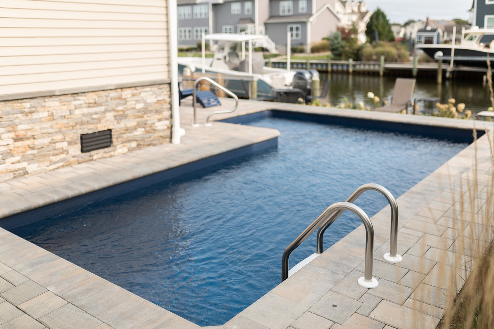 A small rectangular in-ground pool with a ladder, surrounded by a stone and concrete patio, overlooking a marina with boats and houses in the background.