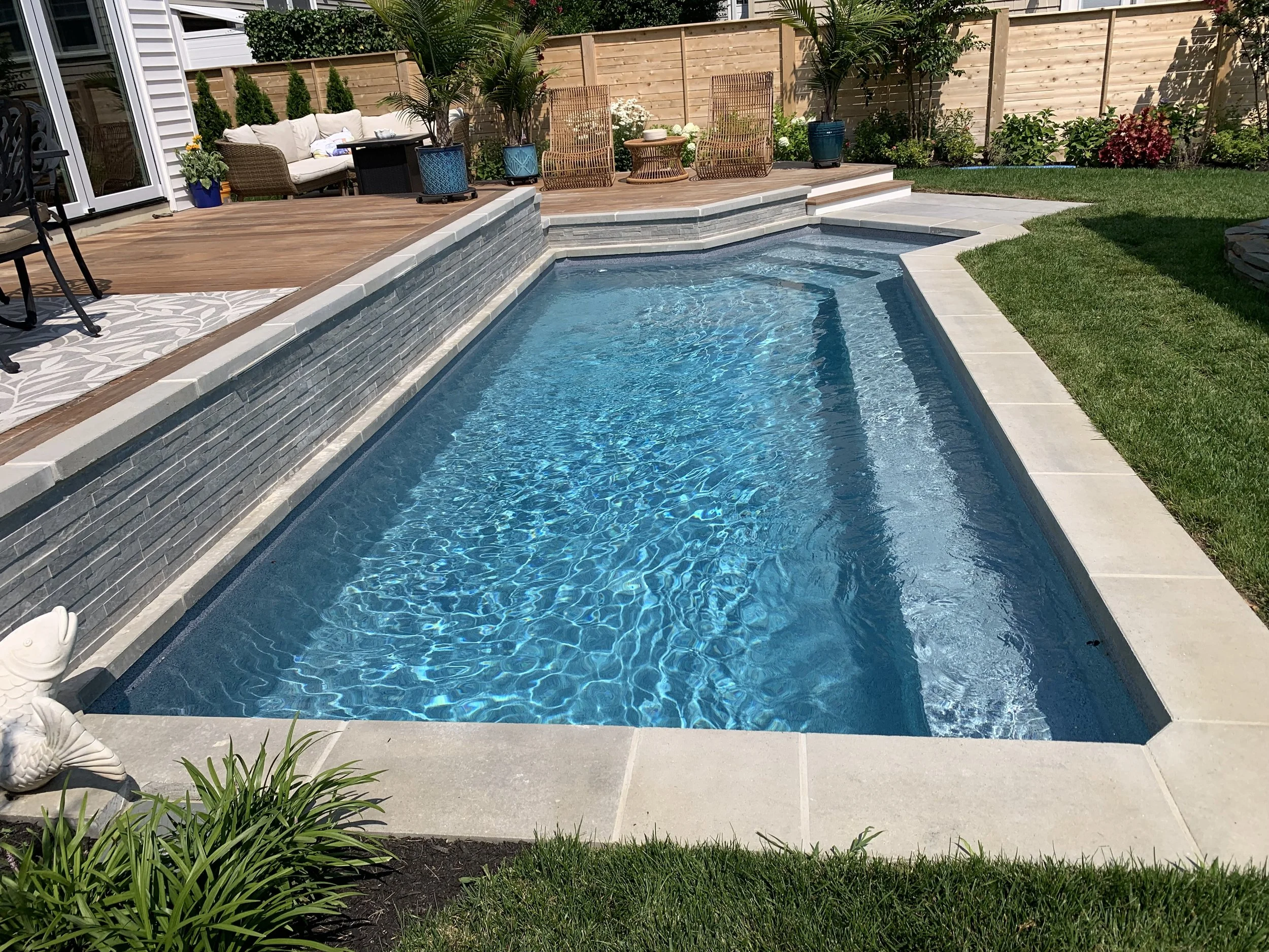 A backyard with a swimming pool, a wooden deck with outdoor furniture, potted plants, and a wooden privacy fence.