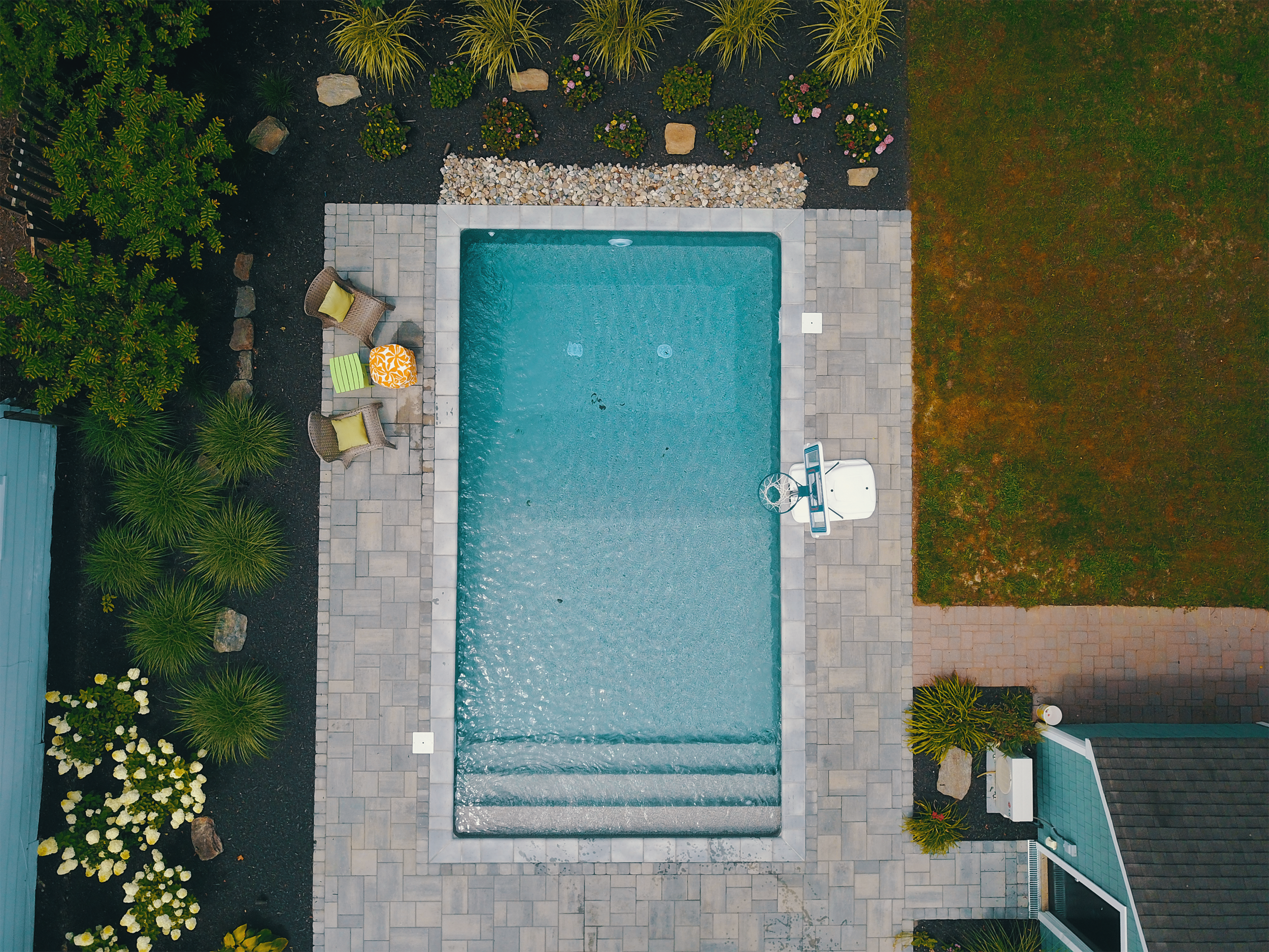 A backyard swimming pool with a basketball hoop on the side, surrounded by pavers, plants, and flowers.