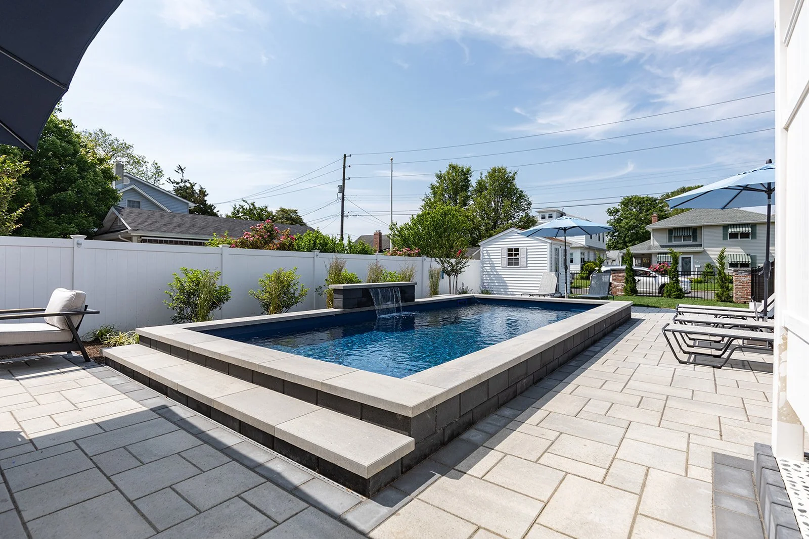 Backyard with swimming pool, patio furniture, umbrellas, and neighboring houses under a blue sky.