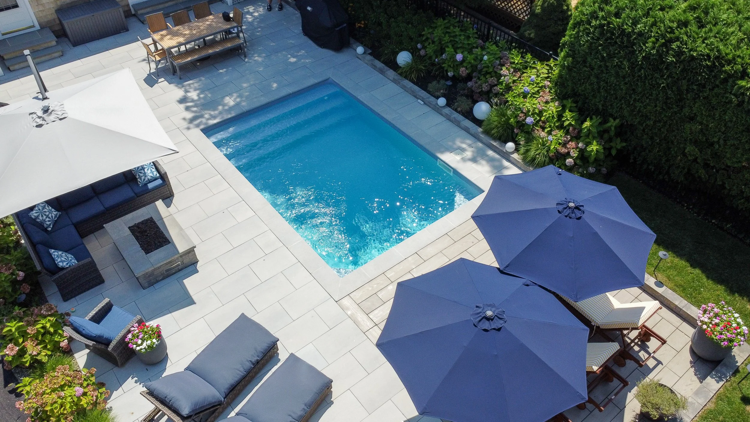 A backyard patio with a swimming pool, outdoor furniture including a sectional sofa, chairs, and tables, shaded by large blue umbrellas, with lush greenery and flowering plants along the border.