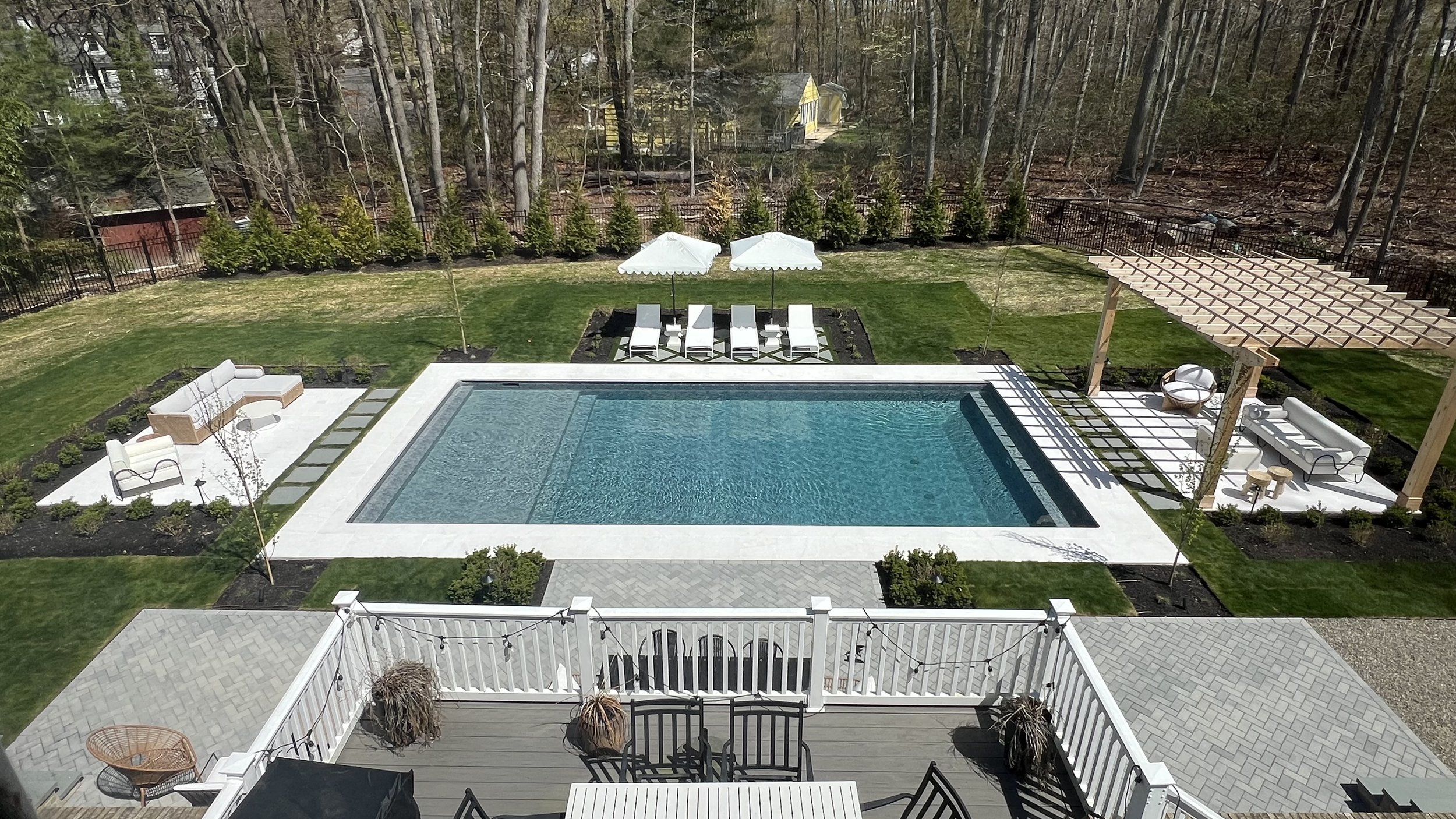 A backyard with a swimming pool surrounded by grass, lounge chairs, umbrellas, and outdoor furniture, enclosed by a black metal fence with trees and houses in the background.