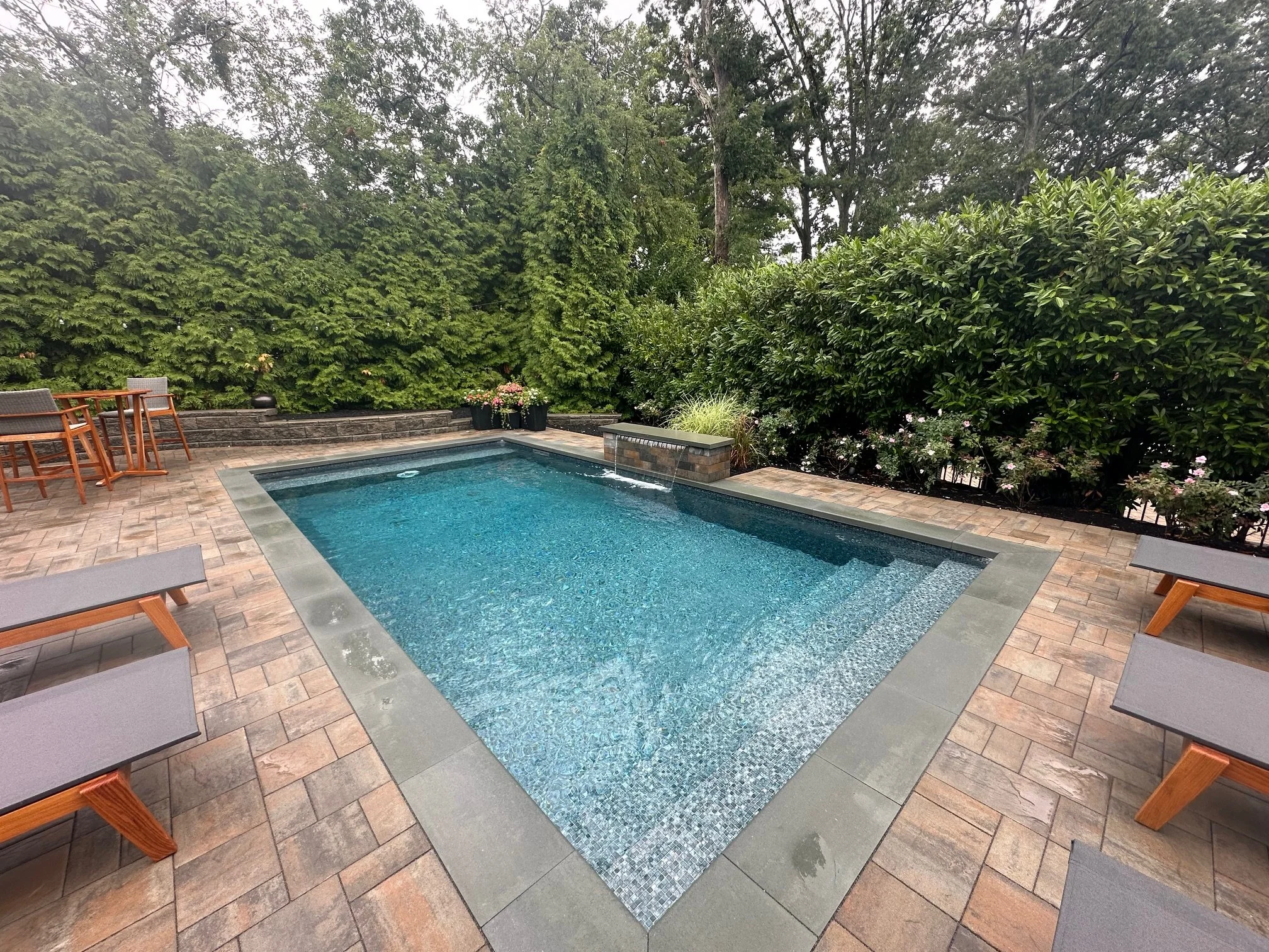 Backyard with a swimming pool, surrounded by brick pavers, wooden chairs, and lush green trees and shrubs.