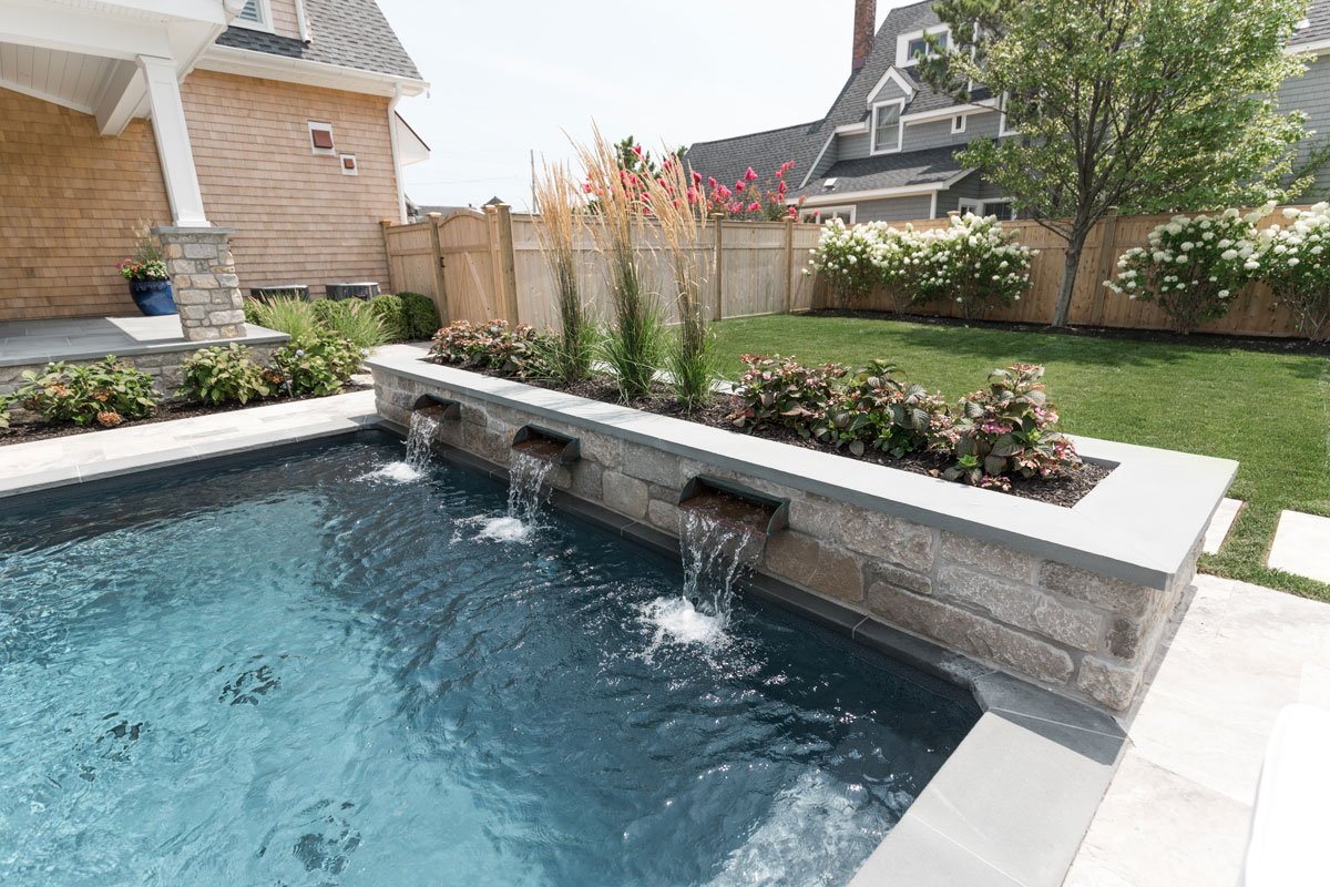 Backyard with a swimming pool, a raised garden bed with flowers, and a lawn with shrubs and trees, surrounded by a wooden fence.