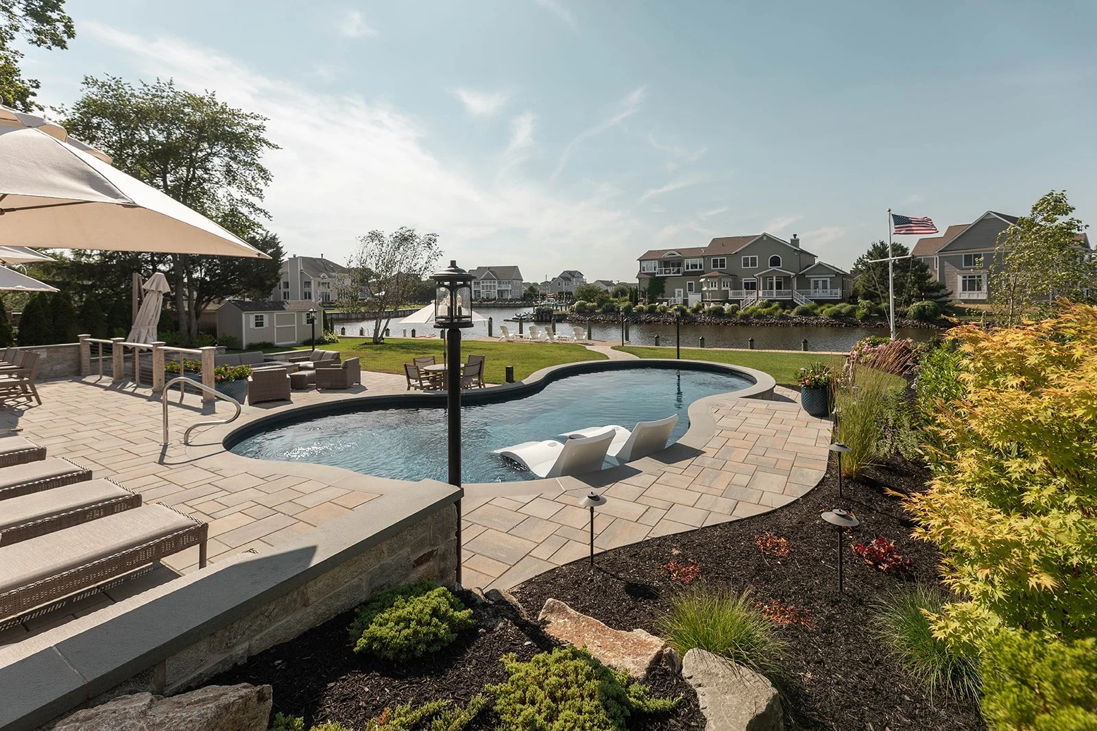A backyard view featuring a kidney-shaped swimming pool with two lounge chairs in the water, outdoor furniture including chairs and umbrellas, a grassy area facing a waterway with houses, and an American flag on a pole amid landscaped plants and tree