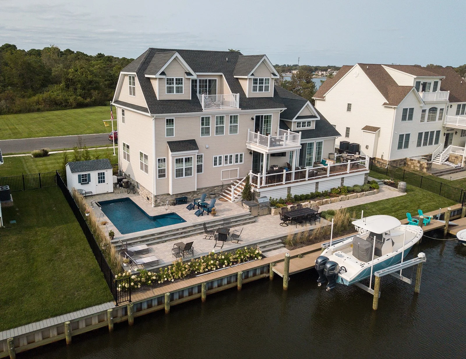 A large, multi-story house by a body of water with a dock and boat. The house has a backyard with a swimming pool, patio furniture, landscaped garden, and balconies.