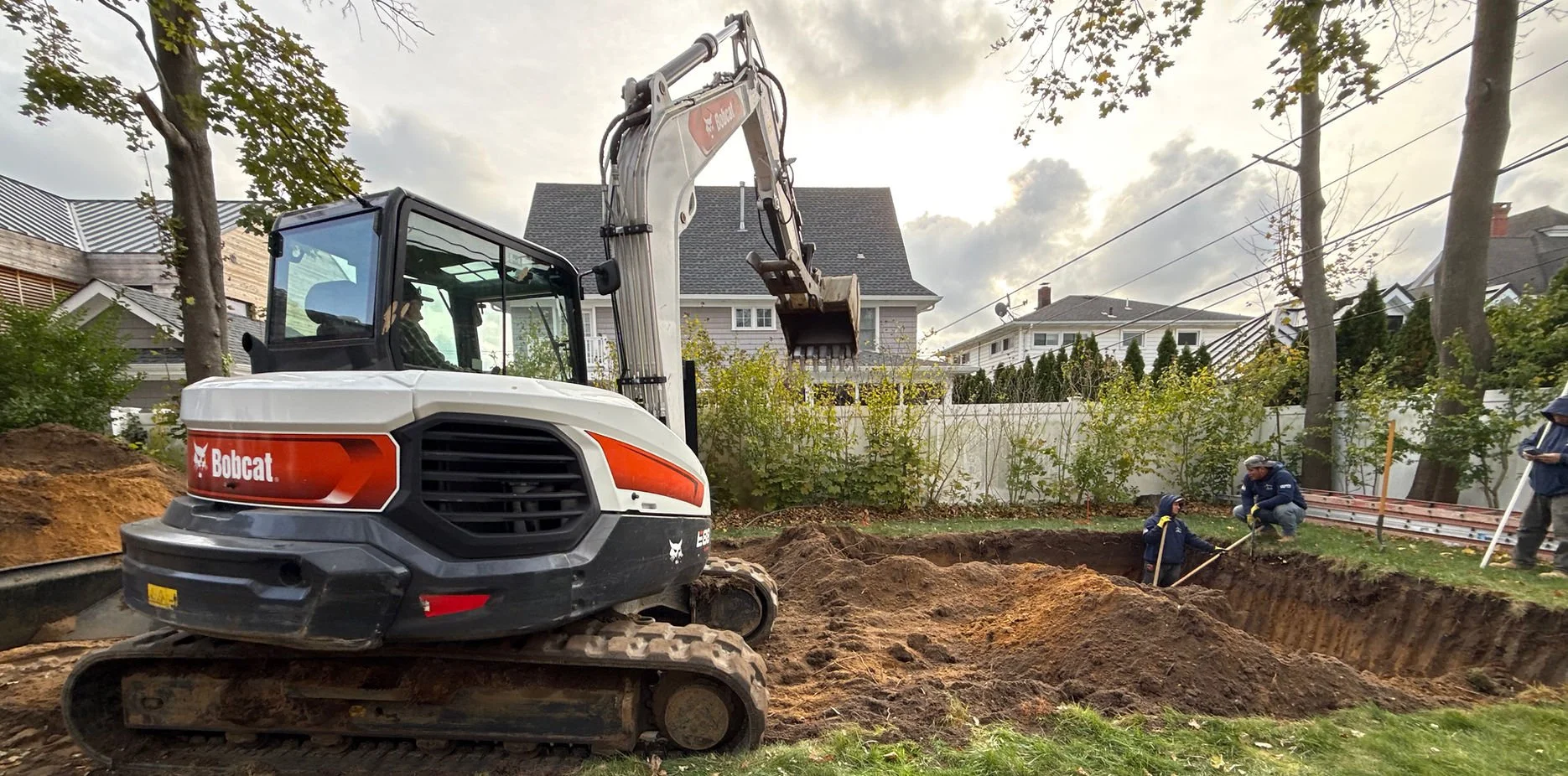 A Bobcat excavator digging a hole in a backyard with two workers measuring and inspecting the site, surrounded by trees and residential houses.