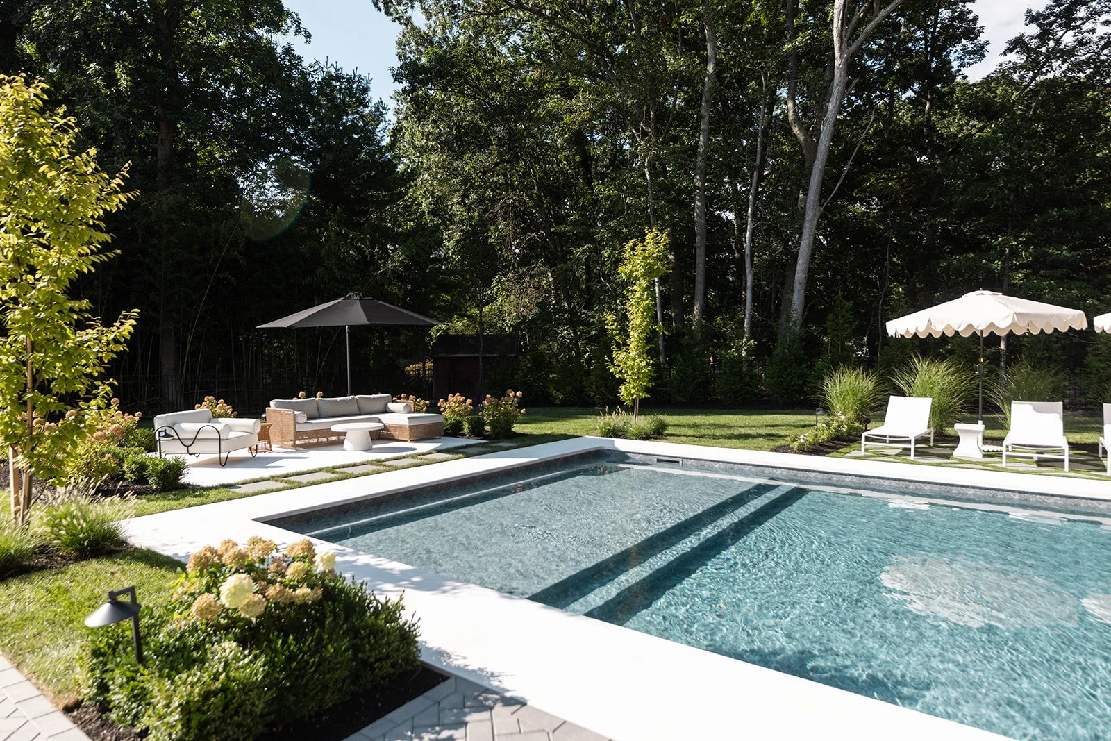 A backyard with a swimming pool, surrounded by outdoor furniture including chairs and umbrellas, lush green trees, and well-maintained landscaping.