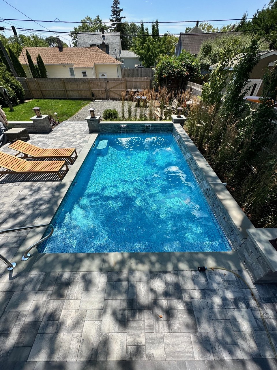 Backyard with a small rectangular swimming pool, wooden lounge chairs, potted plants, a fenced yard, and neighboring houses visible in the background.