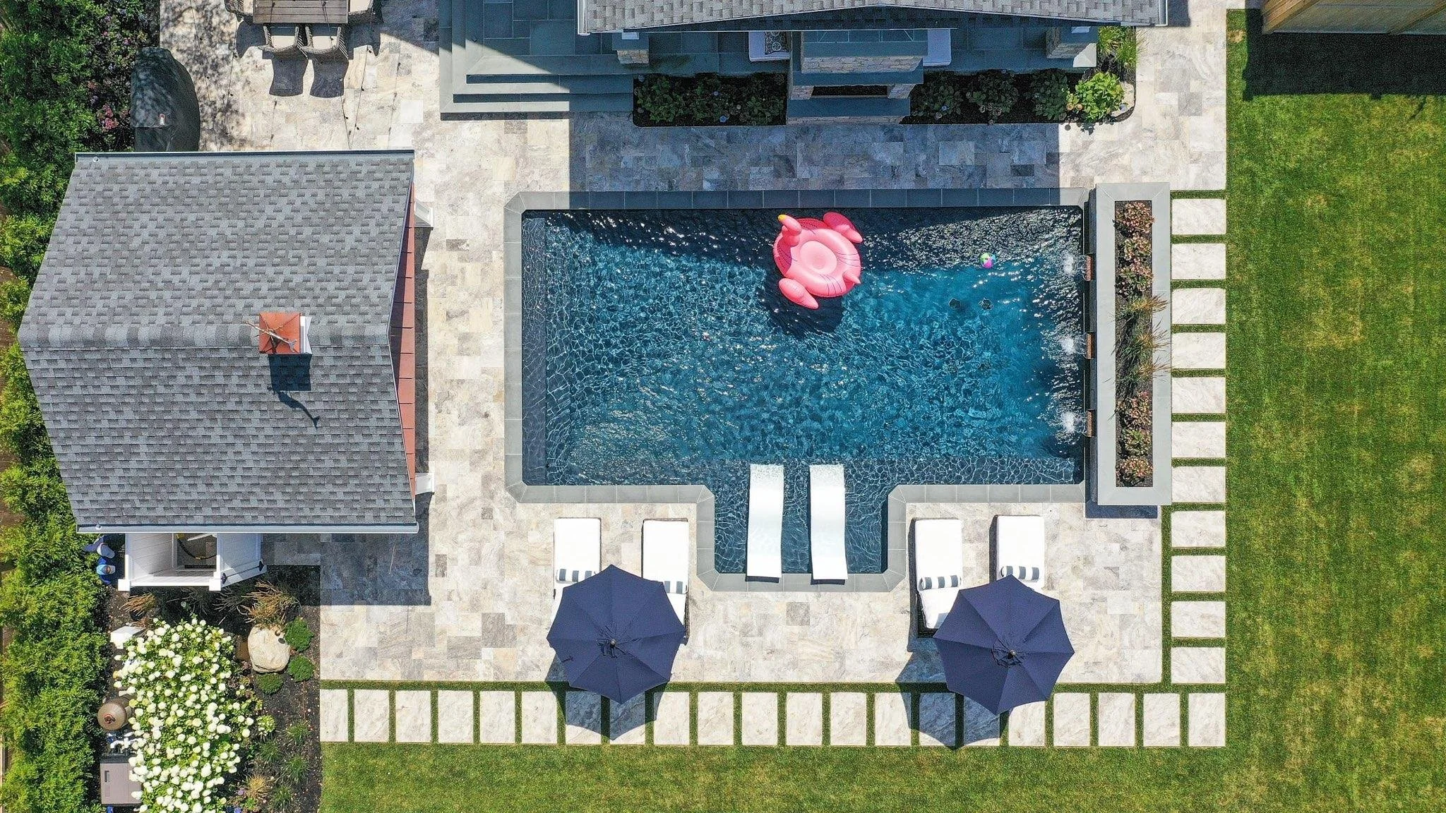 A bird's eye view of a backyard with a swimming pool, pink inflatable unicorn float, and pool toys. Surrounding the pool are patio chairs, umbrellas, a lawn, and garden areas with flowers and shrubs.