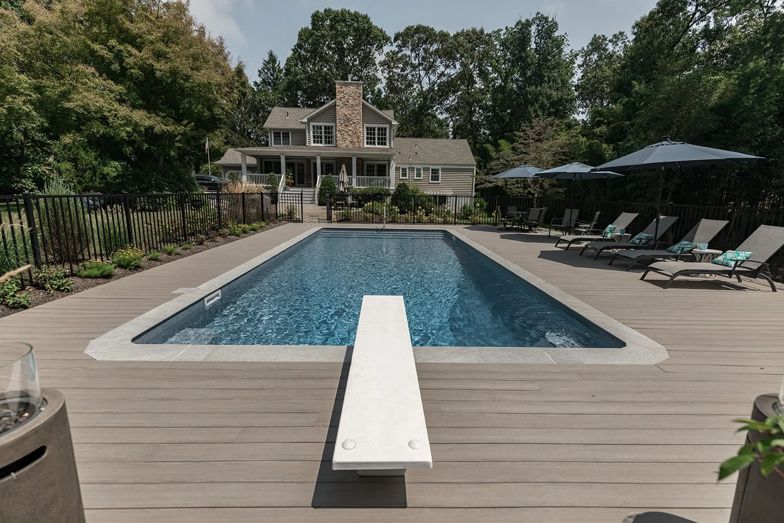 Backyard with a rectangular swimming pool, surrounded by a wooden deck with lounge chairs and umbrellas, and a house with a porch and chimney in the background, all enclosed by a black fence and lush green trees.