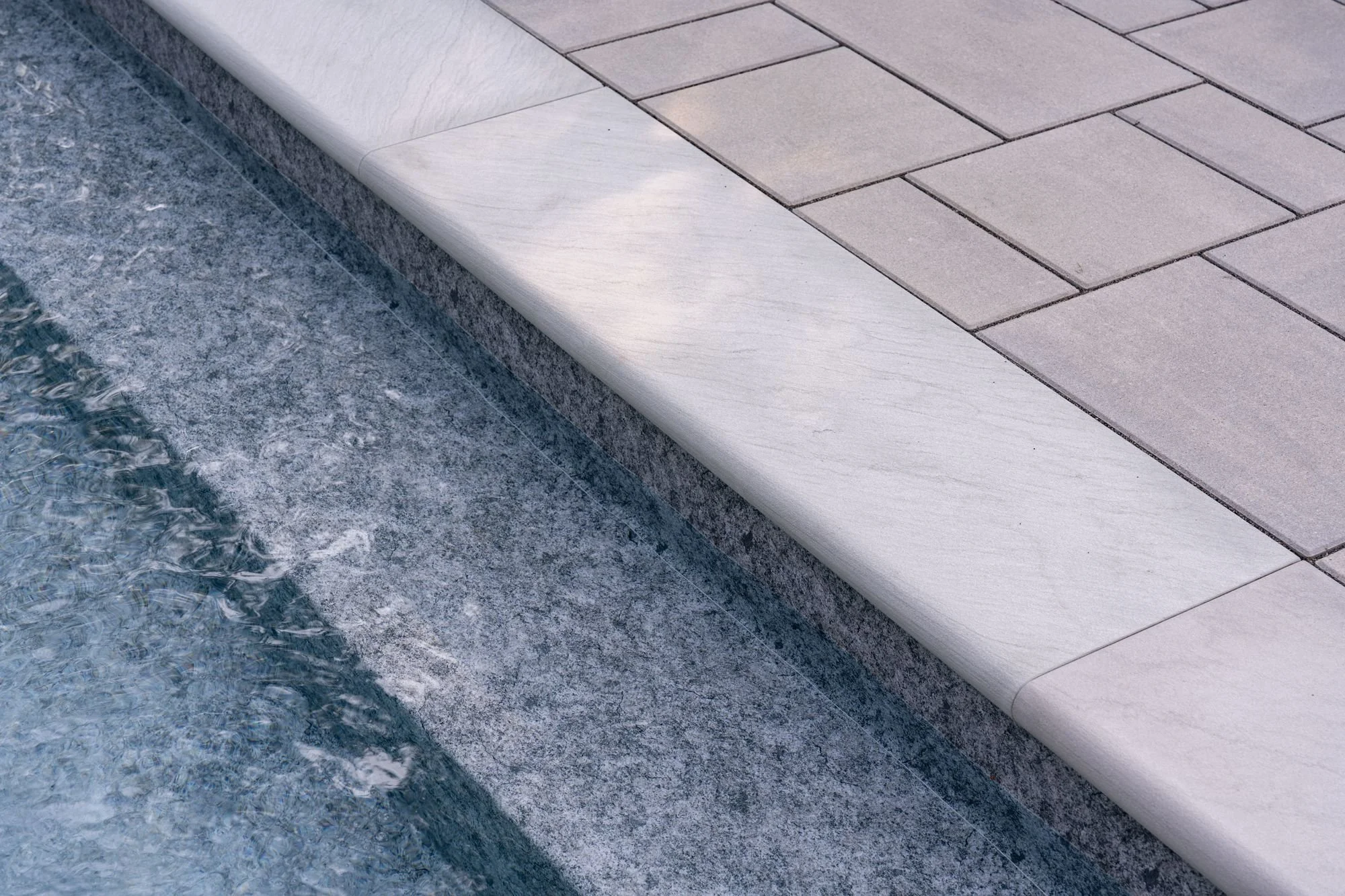 Close-up of a building edge with a smooth, white concrete curb and paving stones made of rectangular tiles, next to a flowing water feature with a textured, dark gray stone surface.