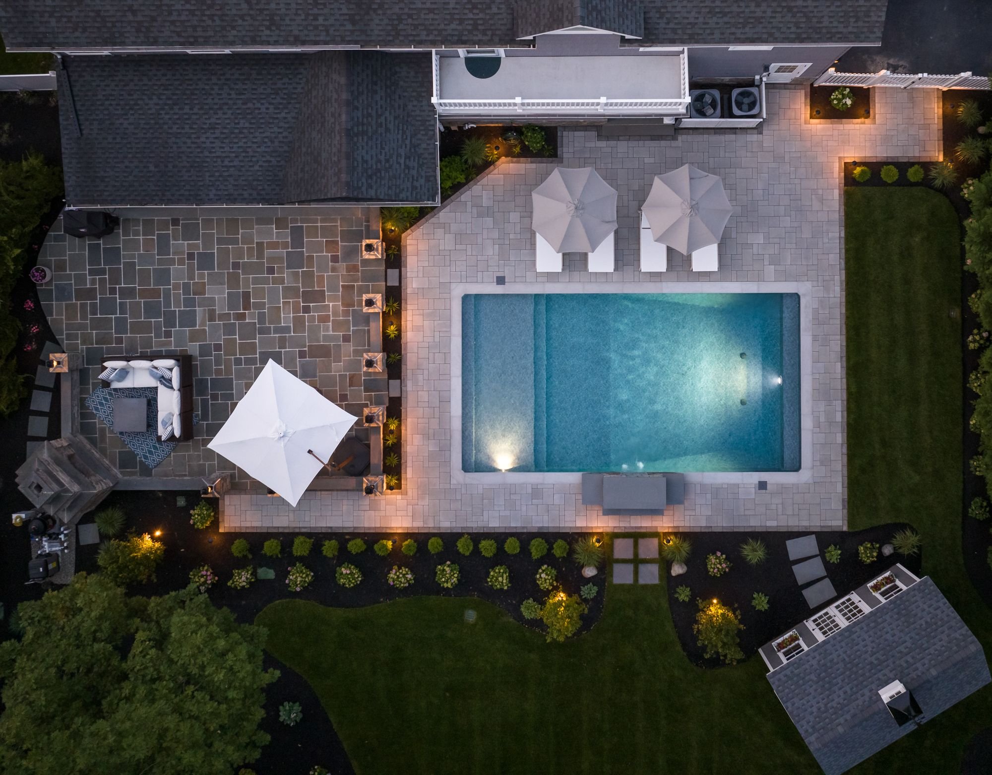 An aerial view of a backyard with a swimming pool, patio furniture with umbrellas, and landscaped gardens at dusk.