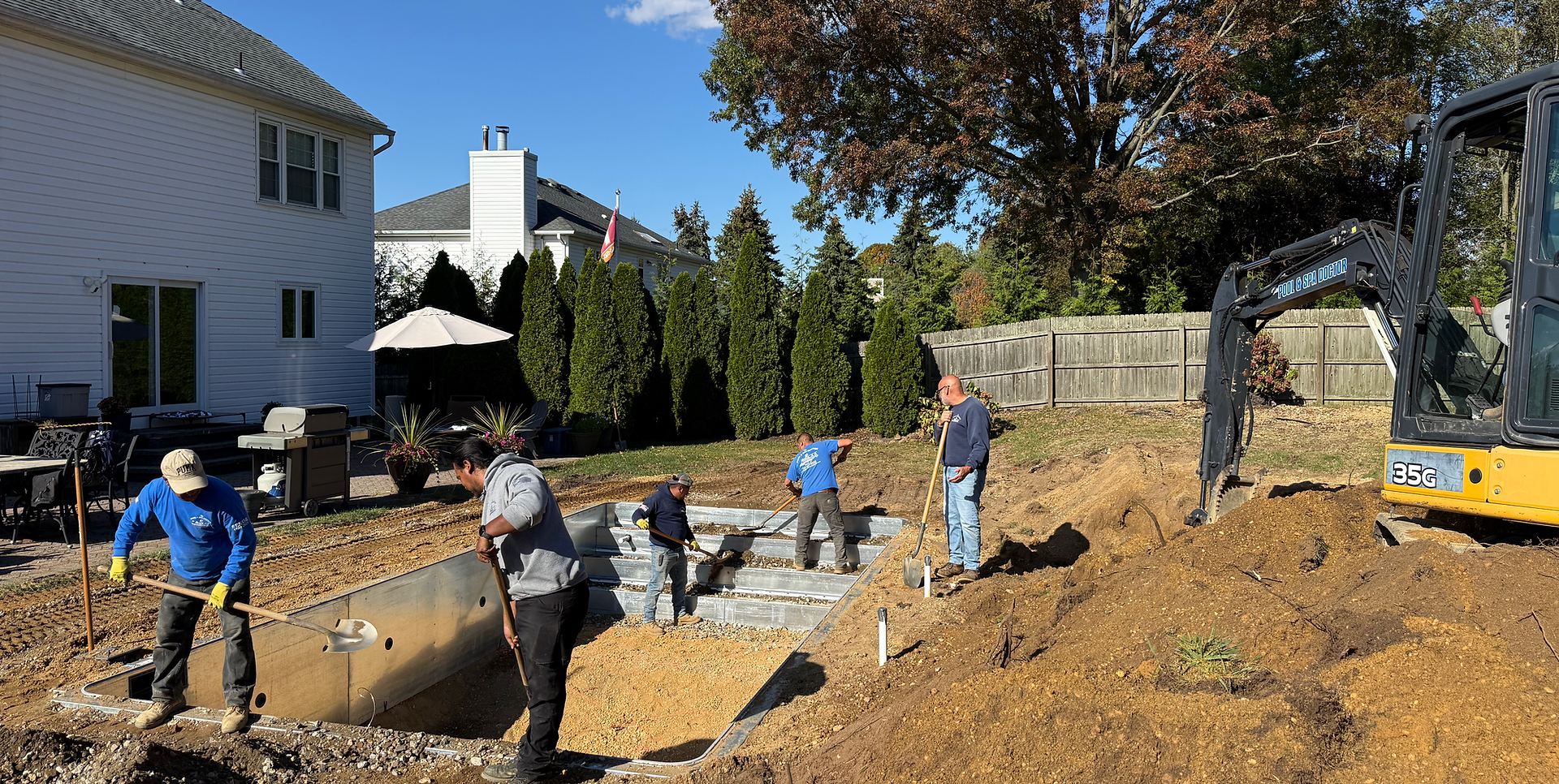Construction workers installing a pool liner in a backyard during daytime, with a house, trees, and construction equipment visible in the background.
