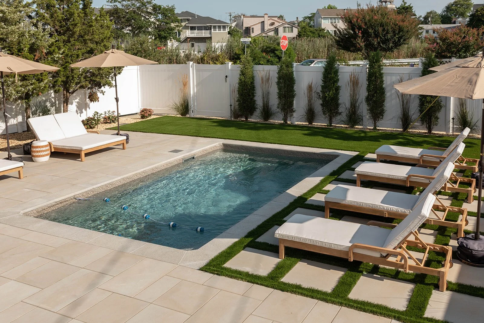 A backyard with a small rectangular swimming pool surrounded by beige paving stones, with four white lounge chairs with cushions and two shaded umbrellas on the right. The yard has a well-manicured grassy area and a white privacy fence with tall, nar
