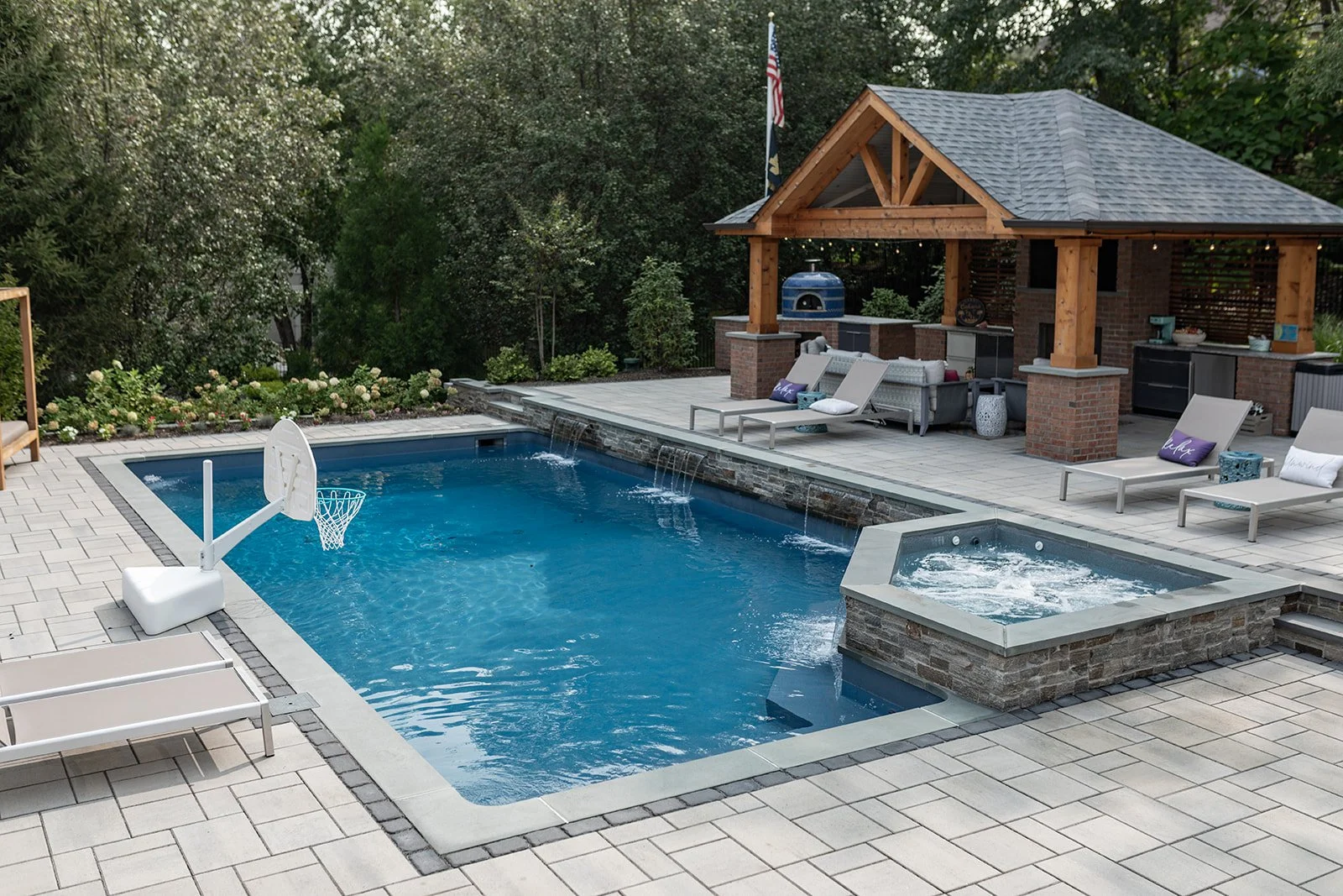 An outdoor backyard with a swimming pool, lounge chairs, hot tub, and a shaded area with outdoor kitchen and seating, surrounded by trees and greenery.