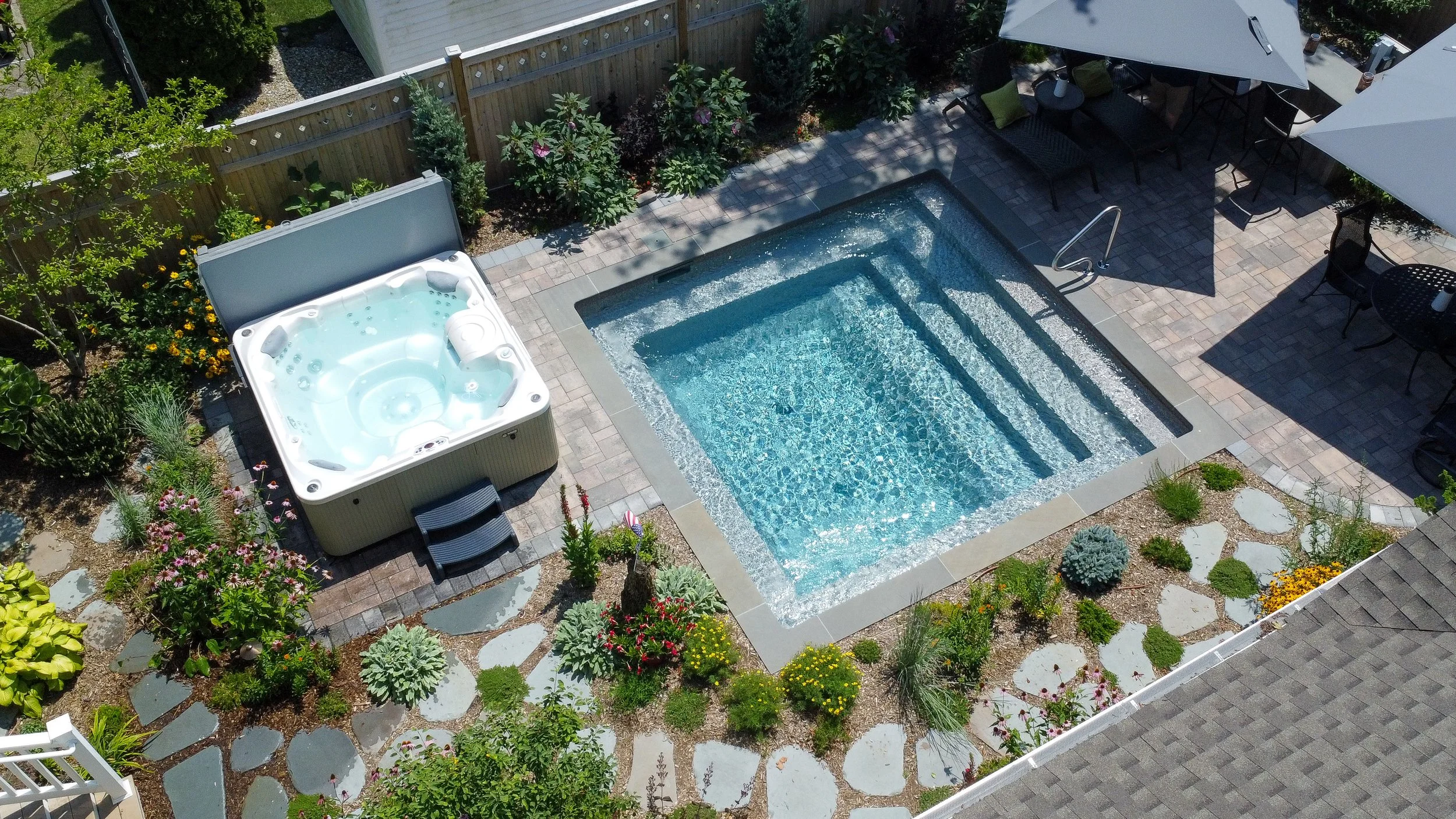 A backyard with a swimming pool, a hot tub, patio tables with umbrellas, and surrounding plants and flowers.
