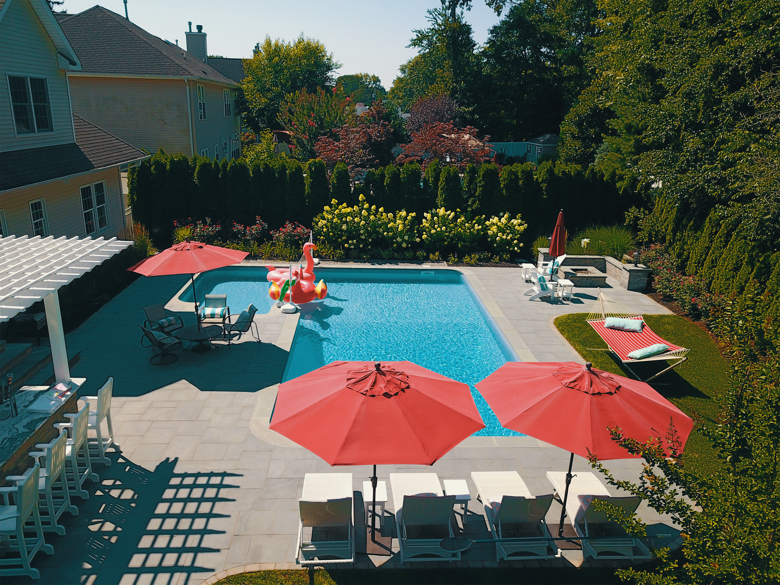 A backyard swimming pool with red umbrellas, a pink flamingo pool float, and outdoor furniture. The area is surrounded by trees and bushes, with houses visible in the background.