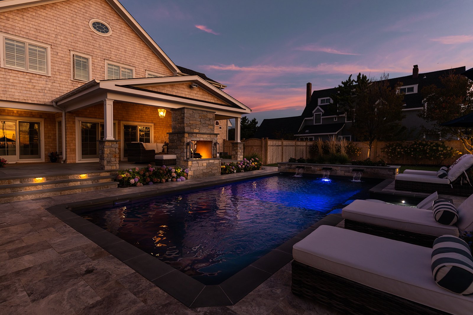 Backyard with lit swimming pool, outdoor fireplace, lounge chairs, and patio area at dusk.