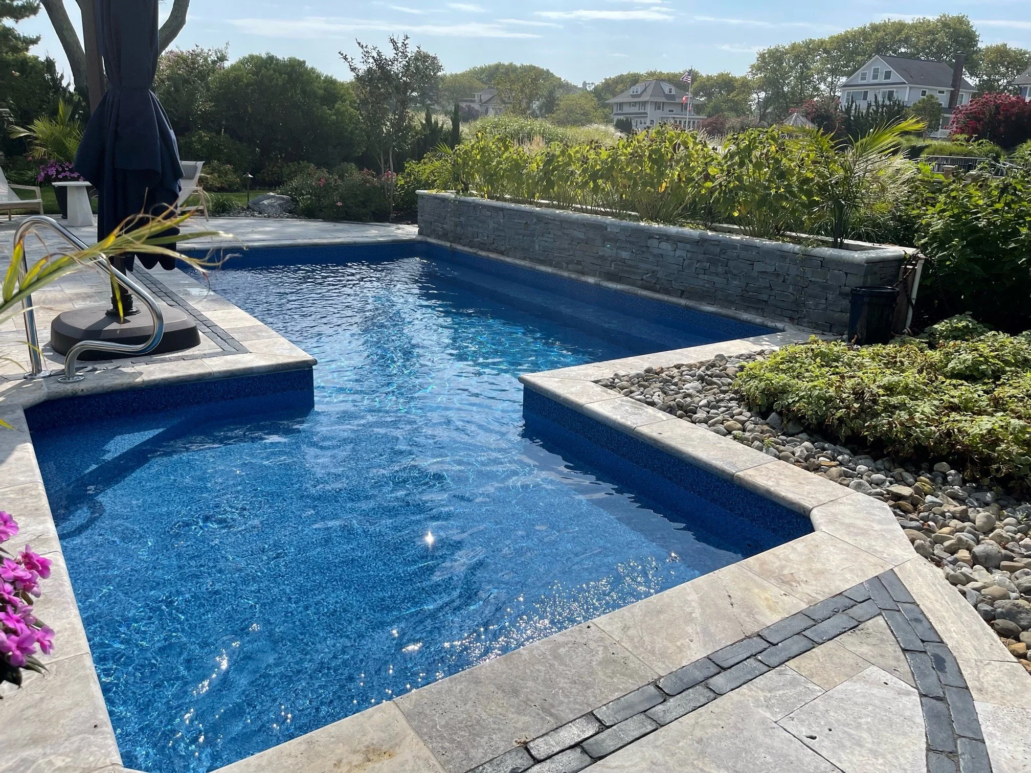 An outdoor swimming pool with clear blue water, surrounded by a stone patio and landscaping with shrubs and rocks. In the background, there are houses and trees under a partly cloudy sky.