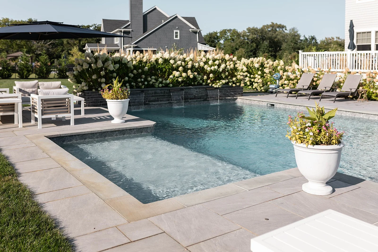 A backyard swimming pool with seating area, large potted flowers, and a background of houses and flowering bushes.