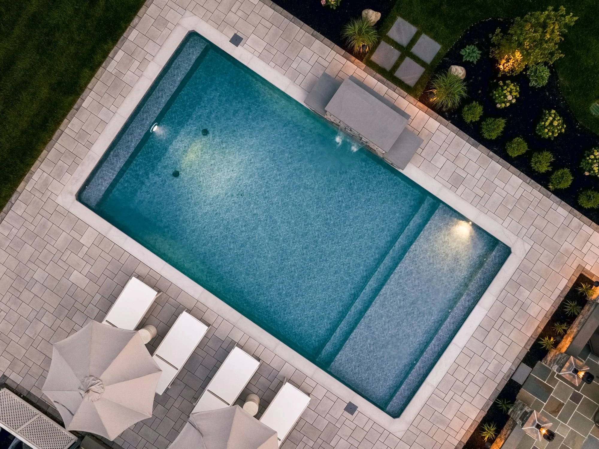 An aerial view of a backyard swimming pool with a patio, patio chairs, umbrellas, and landscaped plants and trees along the edges.