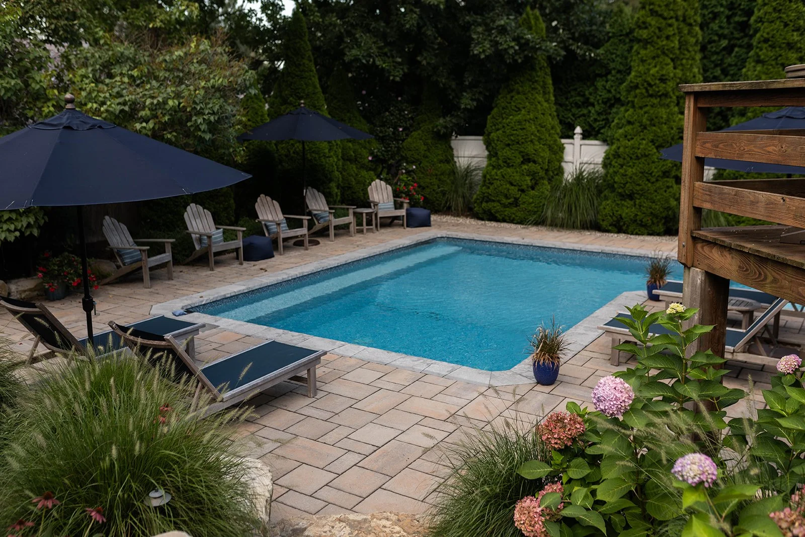 A backyard swimming pool surrounded by beige brick pavers, with Adirondack chairs and umbrellas, lush green trees, and flowering plants.