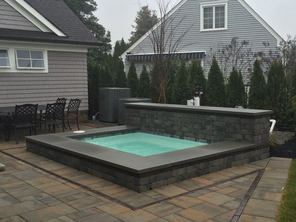Small backyard hot tub with stone and brick surround, porch with patio furniture, and trees and houses in the background.