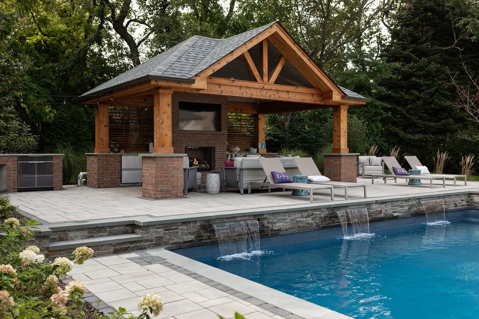 Backyard with a swimming pool, a stone patio, a covered outdoor kitchen and living area with a fireplace, and lounge chairs.