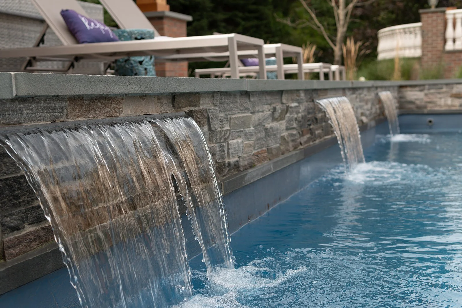 Close-up of a backyard swimming pool with a stone waterfall feature and deck chairs in the background.