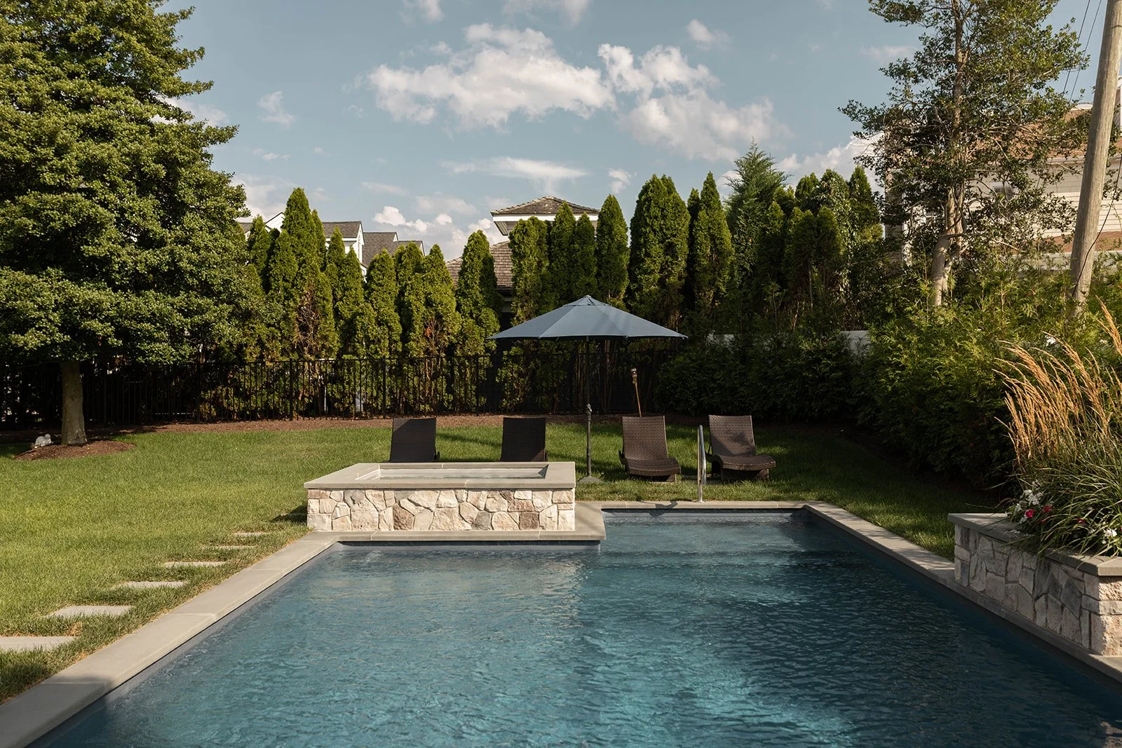 Backyard with swimming pool, lounge chairs, umbrella, lush green trees, and a fence under a partly cloudy sky.